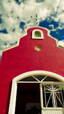 A small, vibrant red chapel with a decorative white trim stands against a backdrop of a bright blue sky with fluffy white clouds. The rooftop features a cross, and a simple entrance with a white door is visible.