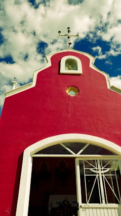 A small, vibrant red chapel with a decorative white trim stands against a backdrop of a bright blue sky with fluffy white clouds. The rooftop features a cross, and a simple entrance with a white door is visible.