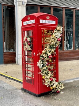 A traditional red telephone booth adorned with festive decorations, including golden leaves, red and gold baubles, and a garland. The booth has the word 'PADDINGTON' displayed at the top. It is situated on a pavement with a modern building in the background.