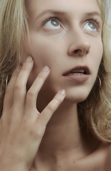 A person with light-colored hair gazes upwards with a thoughtful expression and touching their face with their hand. The facial expression conveys contemplation or wonder, with wide-open eyes and slightly parted lips. The lighting is soft and muted, highlighting the features and skin tone subtly.