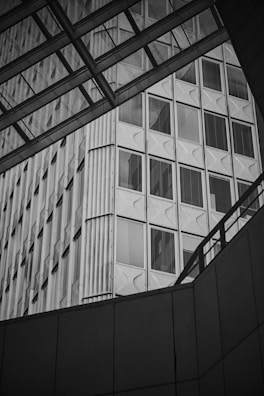 A modern building with a facade featuring large glass windows framed by metallic structures. The architectural design includes geometric patterns and a play of light and shadows, enhanced by the monochrome effect.