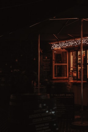 A cozy and dimly lit outdoor food stall with warm string lights hanging above. The stall has a menu board and several signs promoting items such as authentic Cornish pasties. The atmosphere suggests a welcoming and rustic setting.