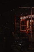 A cozy and dimly lit outdoor food stall with warm string lights hanging above. The stall has a menu board and several signs promoting items such as authentic Cornish pasties. The atmosphere suggests a welcoming and rustic setting.