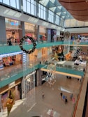 A spacious multi-level shopping mall interior adorned with festive decorations. Large wreaths and golden star ornaments hang from the ceiling. Shoppers walk below on the polished floors, and various stores are visible on both levels.