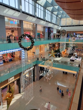 A spacious multi-level shopping mall interior adorned with festive decorations. Large wreaths and golden star ornaments hang from the ceiling. Shoppers walk below on the polished floors, and various stores are visible on both levels.