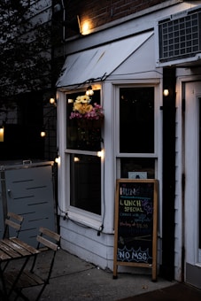 A cozy outdoor dining area with a small table and chairs is in front of a restaurant. The restaurant has a white facade with a hanging basket of yellow and pink flowers near the window. A chalkboard sign advertises a lunch special with text about a spring roll and a price, along with 'No MSG'. String lights are hanging, providing a warm and inviting atmosphere.