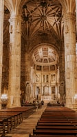 An ornate cathedral interior features tall, detailed columns supporting a vaulted ceiling with intricate ribbed patterns. The central aisle leads to an altar adorned with religious icons and flanked by artwork. Rows of wooden pews are arranged symmetrically.