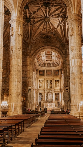 An ornate cathedral interior features tall, detailed columns supporting a vaulted ceiling with intricate ribbed patterns. The central aisle leads to an altar adorned with religious icons and flanked by artwork. Rows of wooden pews are arranged symmetrically.