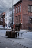 A weathered armchair and a metal walker are placed on the sidewalk of an urban street. In the background, there are several residential buildings with brick and siding facades. The sky is overcast, and parked cars line the street.