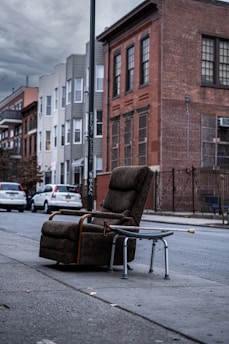 A weathered armchair and a metal walker are placed on the sidewalk of an urban street. In the background, there are several residential buildings with brick and siding facades. The sky is overcast, and parked cars line the street.