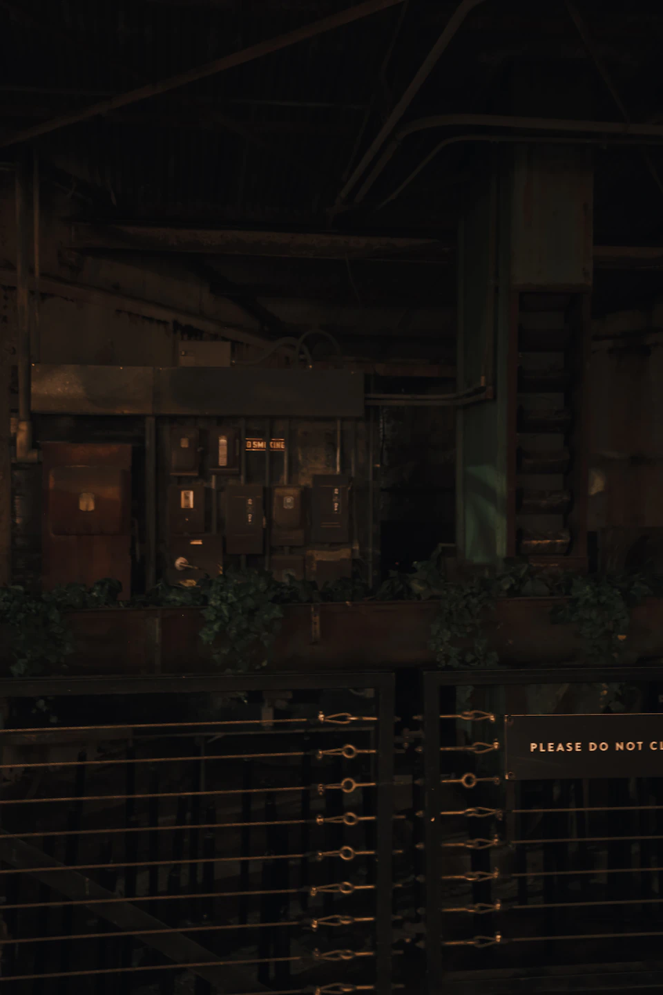 The image depicts a dimly lit room with a metal railing on one side and a wooden fence on the other, both adorned with plants. The railing is made of black metal and features several plants growing from it. On the opposite side of the room, there are two large windows covered by green curtains that allow some light to enter the space.

The walls of the room are painted in a dark color, creating an atmosphere of mystery. A sign on one wall reads "Please do not climb", indicating potential hazards or restricted areas within the building. The floor is made of concrete and features several metal beams running across it, adding to the industrial feel of the space.