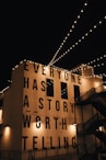 String lights are hung across a night sky, illuminating a building facade with the large, bold text 'EVERYONE HAS A STORY WORTH TELLING'. The building is adorned with industrial-style architecture including staircases and lamps.
