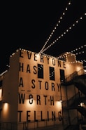 String lights are hung across a night sky, illuminating a building facade with the large, bold text 'EVERYONE HAS A STORY WORTH TELLING'. The building is adorned with industrial-style architecture including staircases and lamps.