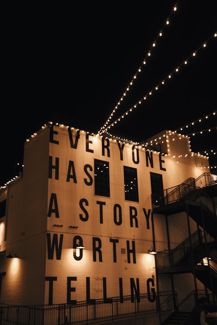 String lights are hung across a night sky, illuminating a building facade with the large, bold text 'EVERYONE HAS A STORY WORTH TELLING'. The building is adorned with industrial-style architecture including staircases and lamps.
