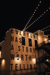 String lights are hung across a night sky, illuminating a building facade with the large, bold text 'EVERYONE HAS A STORY WORTH TELLING'. The building is adorned with industrial-style architecture including staircases and lamps.
