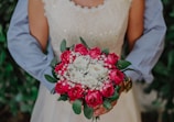 A person in formal attire holds a bouquet of flowers, primarily consisting of red roses and white flowers, surrounded by green leaves. The outfit appears to be bridal or formal, with sequined details visible. The background is out of focus, featuring greenery.