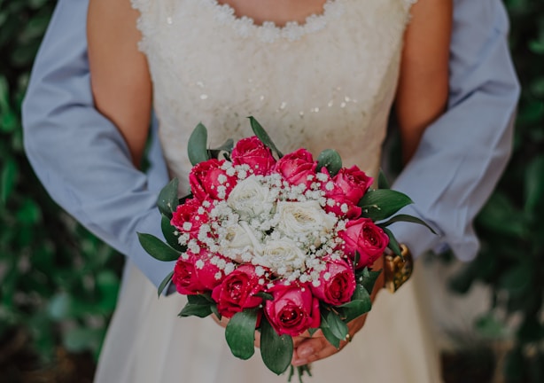 A person in formal attire holds a bouquet of flowers, primarily consisting of red roses and white flowers, surrounded by green leaves. The outfit appears to be bridal or formal, with sequined details visible. The background is out of focus, featuring greenery.