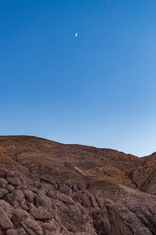 A rugged, rocky landscape with a clear sky above. A small crescent moon is visible in the upper portion of the scene, contrasting with the earthy tones of the terrain.