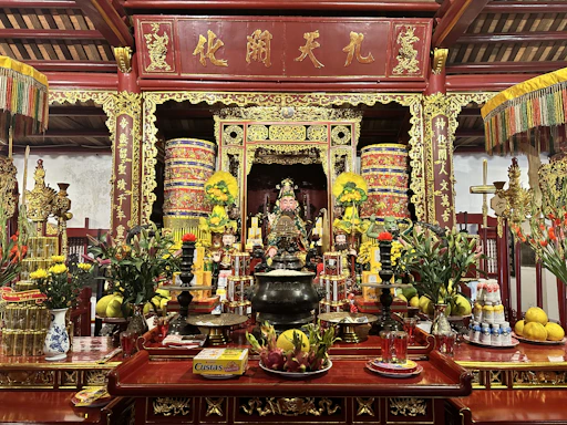 An ornate altar laden with offerings, colorful flowers, and decorative elements, featuring a central figure surrounded by intricate golden carvings and traditional Asian motifs. Candles and incense sticks are visible, adding to the ceremonial ambiance.