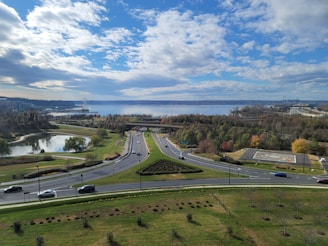 A scenic view of a roadway surrounded by green landscaping, small ponds, and rows of trees under a partly cloudy sky. Vehicles are moving along the roads with a large body of water and urban structures visible in the background.
