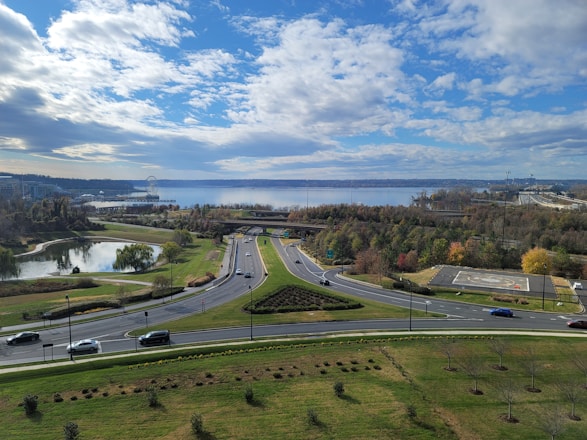 A scenic view of a roadway surrounded by green landscaping, small ponds, and rows of trees under a partly cloudy sky. Vehicles are moving along the roads with a large body of water and urban structures visible in the background.