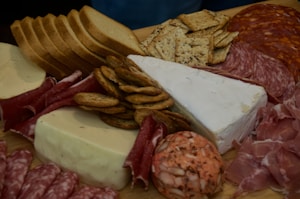 A delicious assortment of charcuterie items including various types of sliced meats like salami and prosciutto, different types of cheese such as brie and blocks of white cheese, and a variety of crackers and sliced bread arranged on a wooden board.