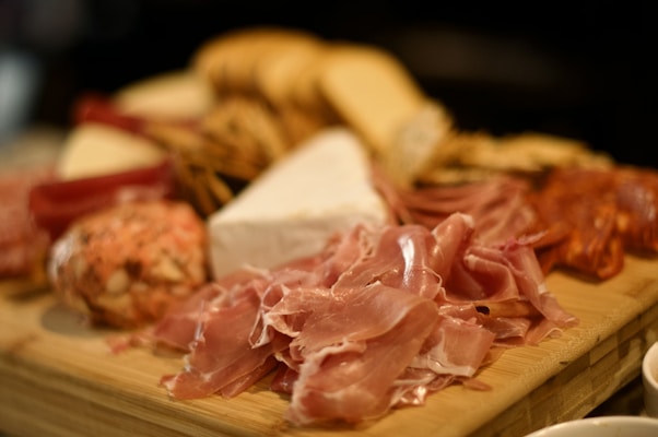 A charcuterie board displays an assortment of cured meats, including slices of prosciutto and salami, accompanied by wedges of cheese, crackers, and breadsticks. The items are neatly arranged on a wooden board, creating a rustic and inviting presentation.