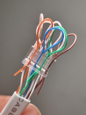 A close-up view of a network cable connector with exposed twisted pair wires, each in different colors including orange, white, green, and blue, emerging from the connector. The connector is transparent, revealing the internal organization of the wires. The background is blurred, highlighting the cable in focus.