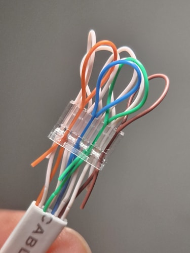 A close-up view of a network cable connector with exposed twisted pair wires, each in different colors including orange, white, green, and blue, emerging from the connector. The connector is transparent, revealing the internal organization of the wires. The background is blurred, highlighting the cable in focus.