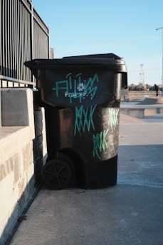 A black trash bin with graffiti is situated on a concrete surface next to a metal fence. The bin has teal-colored graffiti writing on it. In the background, a skate park and some people are visible under a clear blue sky.