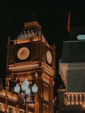 A large clock tower illuminated in warm orange lights stands out against a dark night sky. The architectural details are highlighted, and a group of classic-style street lamps are positioned in the foreground. A building with a slanted roof and the label 'Türkiye' is visible to the right.