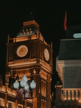 A large clock tower illuminated in warm orange lights stands out against a dark night sky. The architectural details are highlighted, and a group of classic-style street lamps are positioned in the foreground. A building with a slanted roof and the label 'Türkiye' is visible to the right.