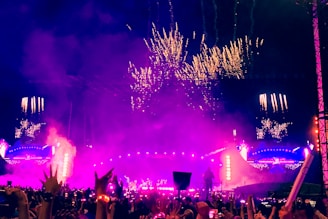 A large, vibrant outdoor concert at night with a crowded audience. The stage is bright with purple and pink lights, and fireworks illuminate the sky above. The atmosphere appears energetic and festive.