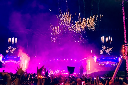 A large, vibrant outdoor concert at night with a crowded audience. The stage is bright with purple and pink lights, and fireworks illuminate the sky above. The atmosphere appears energetic and festive.
