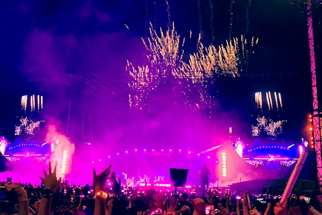 A large, vibrant outdoor concert at night with a crowded audience. The stage is bright with purple and pink lights, and fireworks illuminate the sky above. The atmosphere appears energetic and festive.