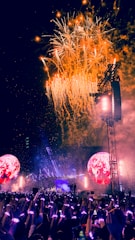 A vibrant concert scene with fireworks exploding in the sky. A large crowd, illuminated by colorful wristbands, stands before a brightly lit stage. Two large circular screens display images, enhancing the atmosphere.