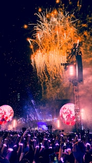 A vibrant concert scene with fireworks exploding in the sky. A large crowd, illuminated by colorful wristbands, stands before a brightly lit stage. Two large circular screens display images, enhancing the atmosphere.