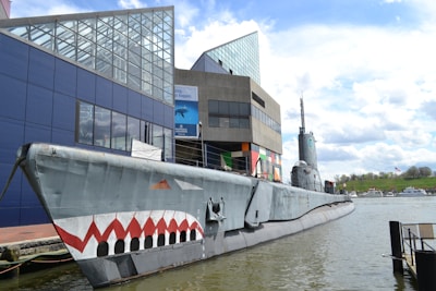 A submarine is docked alongside a modern, angular building with glass and colorful geometric panels. The submarine features a distinctive shark mouth design painted on its bow. The scene is set on a calm body of water with several small boats in the background and a grassy area with a flag visible.
