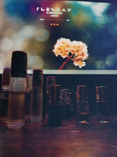 A variety of small glass bottles with different colored liquids are arranged on a wooden surface. In the background, a computer screen displays an image of flowers with a soft, blurred bokeh effect. Text on the screen is visible but not clearly legible.