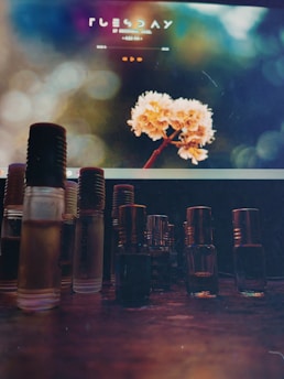 A variety of small glass bottles with different colored liquids are arranged on a wooden surface. In the background, a computer screen displays an image of flowers with a soft, blurred bokeh effect. Text on the screen is visible but not clearly legible.