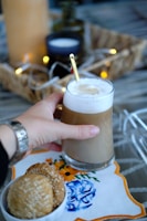A hand is holding a glass of coffee with foam on top. There is a gold-colored spoon resting on the edge of the glass. The setup includes a decorative cloth with blue and orange floral embroidery underneath the glass. Nearby, a bowl contains three sesame cookies. String lights and a wicker basket are blurred in the background, contributing to a cozy ambiance.