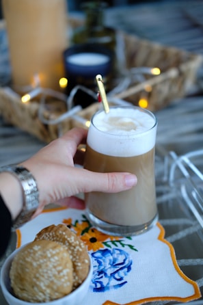 A hand is holding a glass of coffee with foam on top. There is a gold-colored spoon resting on the edge of the glass. The setup includes a decorative cloth with blue and orange floral embroidery underneath the glass. Nearby, a bowl contains three sesame cookies. String lights and a wicker basket are blurred in the background, contributing to a cozy ambiance.