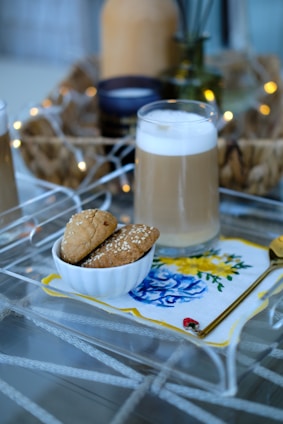 A glass of a milk-based drink with a foamy top is placed on a glass table. Beside it, a white bowl holds several sesame-covered cookies. The setting includes a decorative napkin with blue and yellow floral designs, and a gold spoon rests next to the bowl. Soft, ambient lighting is in the background with a wicker tray containing some glassware adding a cozy atmosphere.