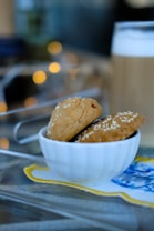 A white bowl containing two sesame-topped cookies is placed on a colorful napkin. In the background, a frothy beverage is visible in a tall glass. Warm bokeh lights add a cozy ambiance.