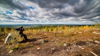 A person wearing a backpack uses a camera to take a photo in a scenic landscape. Surrounded by a rocky terrain with small trees and bushes, the view extends to a large expanse of flatlands and a distant water body. The sky is filled with dramatic, heavy clouds, suggesting a moody atmosphere.