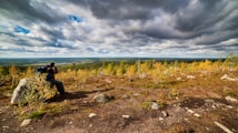 A person wearing a backpack uses a camera to take a photo in a scenic landscape. Surrounded by a rocky terrain with small trees and bushes, the view extends to a large expanse of flatlands and a distant water body. The sky is filled with dramatic, heavy clouds, suggesting a moody atmosphere.