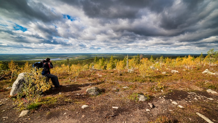A person wearing a backpack uses a camera to take a photo in a scenic landscape. Surrounded by a rocky terrain with small trees and bushes, the view extends to a large expanse of flatlands and a distant water body. The sky is filled with dramatic, heavy clouds, suggesting a moody atmosphere.