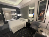 A modern hotel room features a comfortable, neatly arranged bed with white linens. Two contemporary armchairs and a small circular table occupy the corner. A large framed picture of a horse adds character to the room. There is a glass wall with a view into a bathroom, which includes a shower. The room is softly lit by several lamps, and the overall design is sleek and sophisticated.