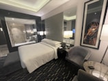 A modern hotel room features a comfortable, neatly arranged bed with white linens. Two contemporary armchairs and a small circular table occupy the corner. A large framed picture of a horse adds character to the room. There is a glass wall with a view into a bathroom, which includes a shower. The room is softly lit by several lamps, and the overall design is sleek and sophisticated.