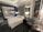 A modern hotel room features a comfortable, neatly arranged bed with white linens. Two contemporary armchairs and a small circular table occupy the corner. A large framed picture of a horse adds character to the room. There is a glass wall with a view into a bathroom, which includes a shower. The room is softly lit by several lamps, and the overall design is sleek and sophisticated.